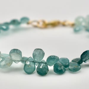 May include: A delicate bracelet featuring teardrop-shaped, faceted blue-green gemstones. The beads are strung together, forming a circular shape, and are connected by a gold-colored clasp. The gemstones vary in shade, creating a gradient effect.