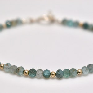 May include: A delicate bracelet featuring small, faceted blue-green gemstones and gold beads. The bracelet is strung on a silver chain.