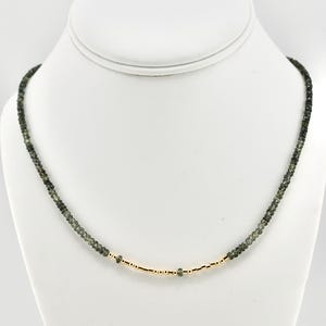 May include: A delicate necklace featuring small, dark green beads and a gold-colored central section. The necklace is displayed on a white mannequin, showcasing its design and craftsmanship. The beads appear to be evenly sized, creating a refined and elegant look.