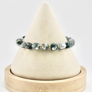 Puede incluir: Una pulsera elástica compuesta por cuentas de ágata musgosa en forma de cubo con patrones jaspeados en verde oscuro, blanco y gris, intercaladas con pequeñas cuentas espaciadoras redondas de metal dorado pulido en secuencia.
