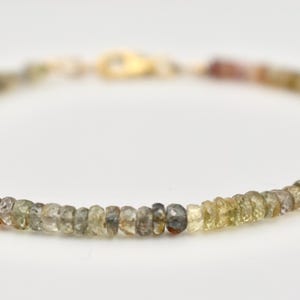 May include: A delicate bracelet featuring small, round beads in shades of brown, green, and yellow. The beads are strung together with a gold-colored clasp. The bracelet is displayed on a white background.
