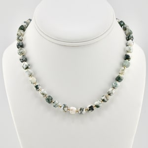 Puede incluir: Un collar con cuentas facetadas y redondeadas que presentan un patrón moteado en blanco y verde oscuro. Las cuentas están separadas por pequeños espaciadores redondos de color oro metálico, creando una secuencia repetitiva a lo largo.