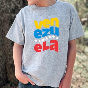 May include: A gray t-shirt with the word "Venezuela" printed on it in yellow, blue, and red letters. The letters are stacked on top of each other, with the word "Venezuela" spelled out in a stylized font.