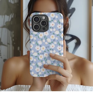 May include: A light blue phone case with a floral pattern. The case features a repeating design of white flowers with green stems and small yellow and red accents. The phone case is held in front of a person.