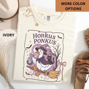 May include: Ivory-colored t-shirt with a graphic of a duck wearing a witch hat and cape, riding a broom. The text "Honkus Ponkus" is above the duck. A black cat sits beside the duck. The shirt has a round neckline.