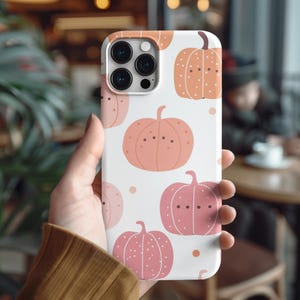 May include: White phone case featuring a repeating pattern of cartoon pumpkins in shades of pink and orange. The pumpkins have dot eyes and are set against a white background. The case has a camera cutout and is held in a person's hand.