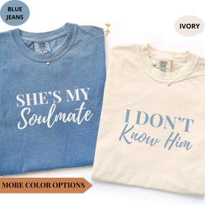 Puede incluir: Dos camisetas, una azul y otra marfil, con texto. La camiseta azul dice "She's My Soulmate" en blanco, y la camiseta marfil dice "I Don't Know Him" en azul. Ambas camisetas tienen un pequeño collar dorado.