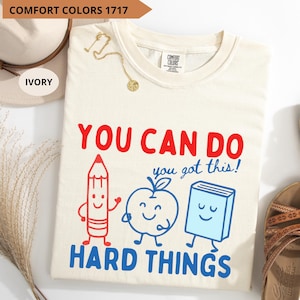 May include: An ivory Comfort Colors 1717 t-shirt featuring a graphic with a red pencil, a blue apple, and a blue book. The text reads YOU CAN DO you got this! HARD THINGS in red and blue lettering. The shirt has a crew neckline and short sleeves.