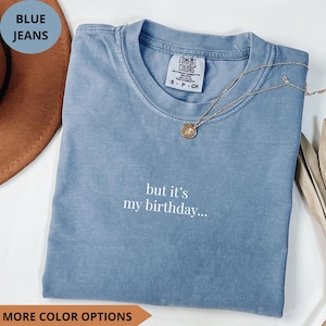 Puede incluir: Una camiseta azul Comfort Colors con el texto "but it's my birthday...". Un collar dorado con un colgante descansa sobre la camiseta. También se ven las palabras "BLUE JEANS" y "MORE COLOR OPTIONS".