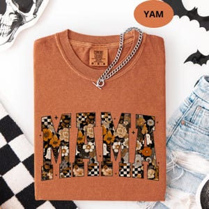 May include: A yam-colored Comfort Colors t-shirt with the word "MAMA" in a floral and checkered design. A silver chain necklace is draped across the shirt. The shirt is folded and placed on a white surface.