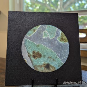 May include: Square black canvas art with a circular abstract design in shades of blue, green, and brown. The artwork is mounted on a black easel. The artist's signature "Erickson, 24" is visible in the lower right corner.