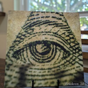 May include: A close-up of a black and white eye-shaped design on a canvas print. The design is made up of concentric circles and lines, creating a hypnotic effect. The canvas is mounted on a black easel with the artist's signature "Erickson, 24" in the bottom right corner.