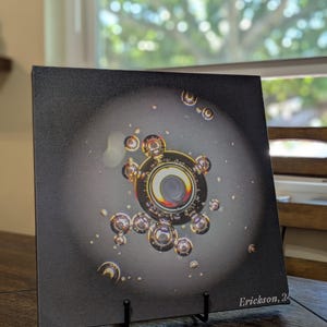 May include: A square canvas print featuring a detailed, abstract design with a central circular element surrounded by smaller, spherical shapes. The artwork has a dark background with a spotlight effect, and the artist's signature is visible.
