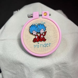 Puede incluir: Un aro de bordado rosa con un fondo de tela blanca. La tela tiene las palabras "needle minder" bordadas. Un alfiler de metal con una imagen de dibujos animados de dos personajes de pelo azul con atuendos rojos está sujeto a la tela.