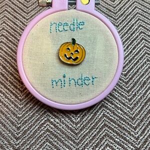 Puede incluir: Un aro de bordado rosa con tela blanca en el interior. La tela tiene las palabras "needle minder" bordadas en hilo azul. Un pequeño protector de aguja con forma de calabaza naranja se encuentra en el centro del aro.