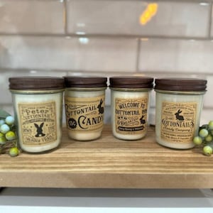 May include: Four glass candle jars with brown lids and labels. The labels feature a bunny rabbit and text that reads "Peter Cottontail", "Cottontail Candy", "Welcome to Cottontail Inn Bed & Breakfast", and "Cottontails Candy Company".
