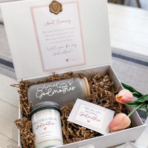 May include: A white gift box with a brown paper filler. The box contains a glass jar with the text "Amelia's Godmother" on it, a candle with the text "Smells like a special Godmother" and a card with the text "Pairs well with becoming Godmother".