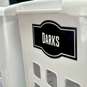 May include: Two white plastic laundry baskets with black labels. The label on the right basket says "DARKS".
