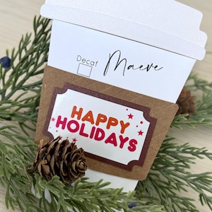 May include: A white paper coffee cup with a brown paper sleeve. The sleeve has a sticker that says "Happy Holidays" in red and orange letters. The cup has a white lid and a sticker that says "Decaf" and "Maeve".