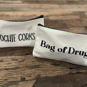May include: Two white canvas zipper pouches with black text. One reads "COOCHIE CORKS" and the other reads "Bag of Drugs". The pouches have black zippers and are on a wooden surface.