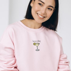 Pu&ograve; includere: Una felpa rosa con la scritta "lemon drop" e un'immagine di un bicchiere da martini con una fetta di limone.