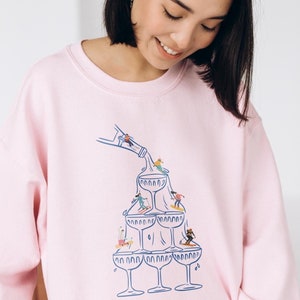 May include: A pink sweatshirt with a graphic of a champagne bottle pouring into a stack of champagne glasses.  People are skiing and snowboarding down the glasses.