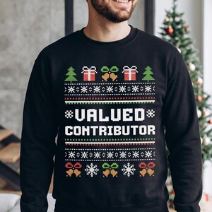 May include: A black long-sleeve sweatshirt featuring a festive holiday-themed pattern in white, red, and green. The design includes rows of Christmas trees, gift boxes, bells, and snowflakes, with the text VALUED CONTRIBUTOR in the center.