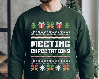 May include: A dark green long-sleeve sweatshirt featuring a white, red, and green pixelated holiday pattern. The design includes Christmas trees, gift boxes, bells, and snowflakes, with the text "MEETING EXPECTATIONS" centered in white.