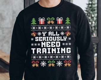May include: A black long-sleeve crewneck sweatshirt featuring a white, red, and green holiday-themed graphic print. The design includes snowflakes, Christmas trees, gift boxes, and bells with the text Y' ALL SERIOUSLY NEED TRAINING in white.