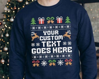 May include: A navy blue long-sleeve sweatshirt featuring a white, red, green, and gold holiday-themed knit pattern. The center displays the text "YOUR CUSTOM TEXT GOES HERE" surrounded by reindeer, gift boxes, bells, and snowflake motifs.