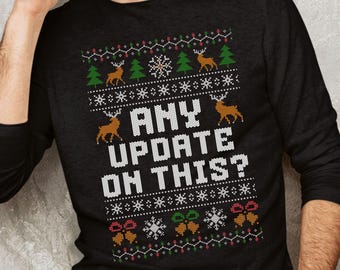 Op de afbeelding: Zwart shirt met lange mouwen met een feestelijk ontwerp. Het shirt heeft een patroon van rendieren, sneeuwvlokken en kerstbomen in groen, rood en wit. De tekst "ANY UPDATE ON THIS?" wordt weergegeven in witte blokletters.
