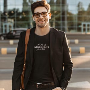 Pu&ograve; includere: Un uomo indossa un blazer nero sopra una t-shirt nera con il testo "NOT A MORNING person" in bianco. Sta sorridendo e guardando la fotocamera.