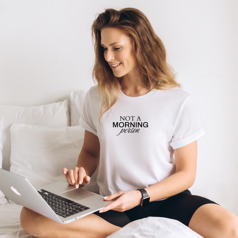 Pu&ograve; includere: Una maglietta bianca con la scritta "NOT A MORNING PERSON" stampata sul davanti. La persona indossa pantaloncini neri e siede su un letto, usando un laptop.
