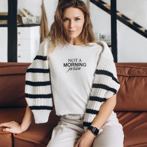 Pu&ograve; includere: Una maglietta bianca con testo nero che recita "NOT A MORNING person". La persona che indossa la maglietta &egrave; seduta su un divano marrone.