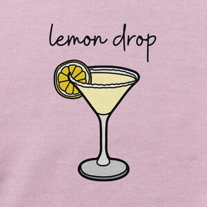Pu&ograve; includere: Una maglietta rosa con il testo "lemon drop" e un'illustrazione di un cartone animato di una fetta di limone e un bicchiere da martini.