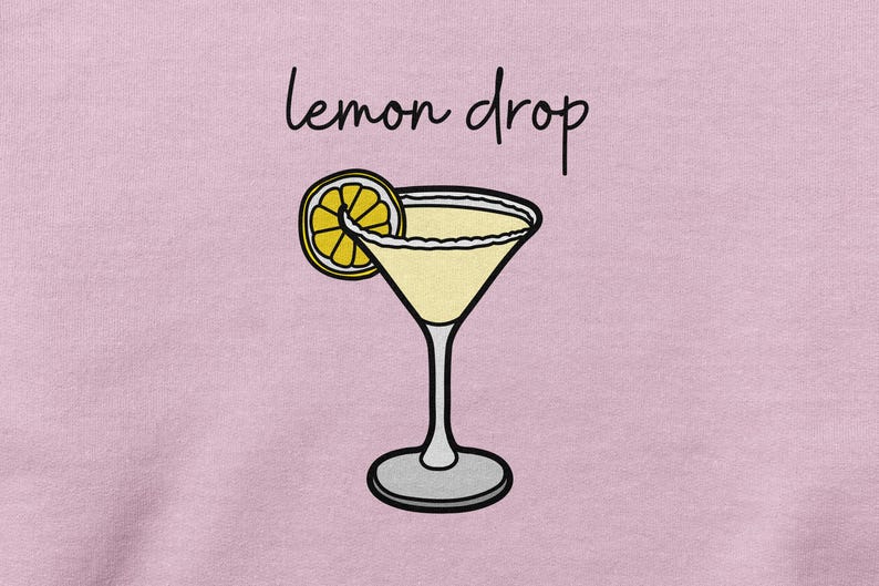 Pu&ograve; includere: Una maglietta rosa con il testo "lemon drop" e un'illustrazione di un cartone animato di una fetta di limone e un bicchiere da martini.