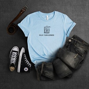 May include: A light blue t-shirt with a graphic of an old fashioned cocktail and the text "Old Fashioned". The shirt is laid out on a gray surface with a pair of black sneakers and a brown leather belt.