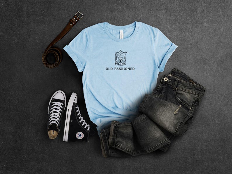 May include: A light blue t-shirt with a graphic of an old fashioned cocktail and the text "Old Fashioned". The shirt is laid out on a gray surface with a pair of black sneakers and a brown leather belt.