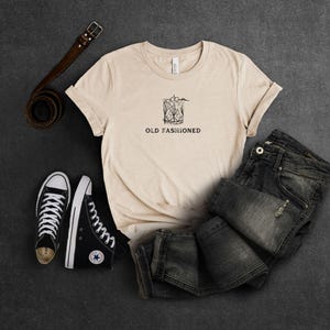 May include: A beige t-shirt with a black and white illustration of an old fashioned cocktail and the text "OLD FASHIONED". The shirt is paired with black sneakers and dark wash jeans.
