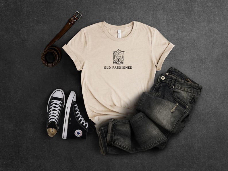 May include: A beige t-shirt with a black and white illustration of an old fashioned cocktail and the text "OLD FASHIONED". The shirt is paired with black sneakers and dark wash jeans.