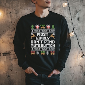 May include: A black long-sleeve sweatshirt featuring a white graphic print with the text MOST LIKELY CAN'T FIND MUTE BUTTON. The design includes pixelated holiday motifs such as green pine trees, red gift boxes, gold bells, and white snowflakes.