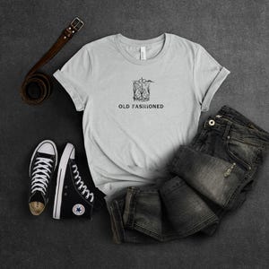 May include: A gray t-shirt with a black and white illustration of an old fashioned cocktail and the text "OLD FASHIONED". The shirt is laid out on a gray surface with a pair of black sneakers and a brown leather belt.