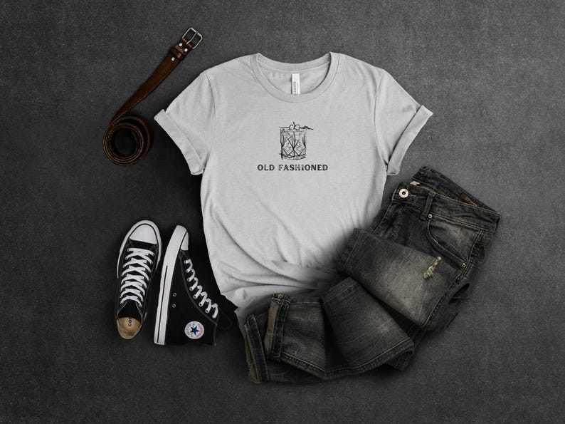 May include: A gray t-shirt with a black and white illustration of an old fashioned cocktail and the text "OLD FASHIONED". The shirt is laid out on a gray surface with a pair of black sneakers and a brown leather belt.
