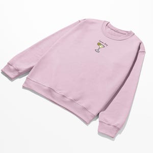 Pu&ograve; includere: Una felpa rosa chiaro con un piccolo disegno ricamato di un bicchiere da cocktail con una fetta di limone e il testo "lemon drop".