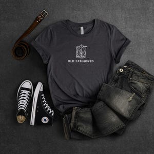 May include: A dark gray t-shirt with a white graphic of a cocktail glass and the text "Old Fashioned". The shirt is laid out on a gray surface with a pair of black sneakers and a brown leather belt.