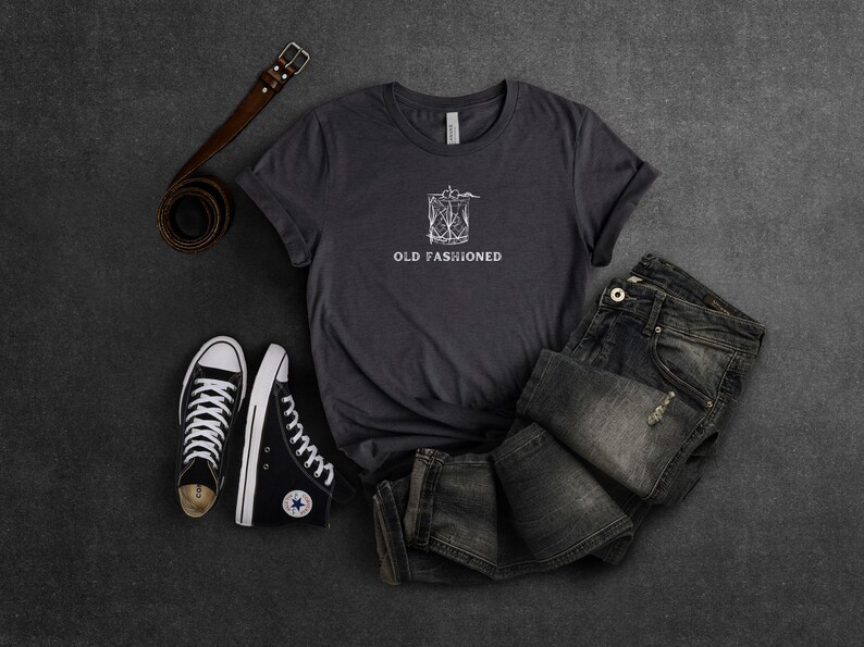 May include: A dark gray t-shirt with a white graphic of a cocktail glass and the text "Old Fashioned". The shirt is laid out on a gray surface with a pair of black sneakers and a brown leather belt.
