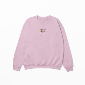 Pu&ograve; includere: Una felpa rosa chiaro con una grafica di un cocktail lemon drop e il testo "lemon drop" sul davanti.