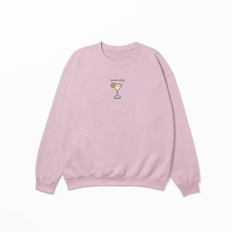 Pu&ograve; includere: Una felpa rosa chiaro con una grafica di un cocktail lemon drop e il testo "lemon drop" sul davanti.