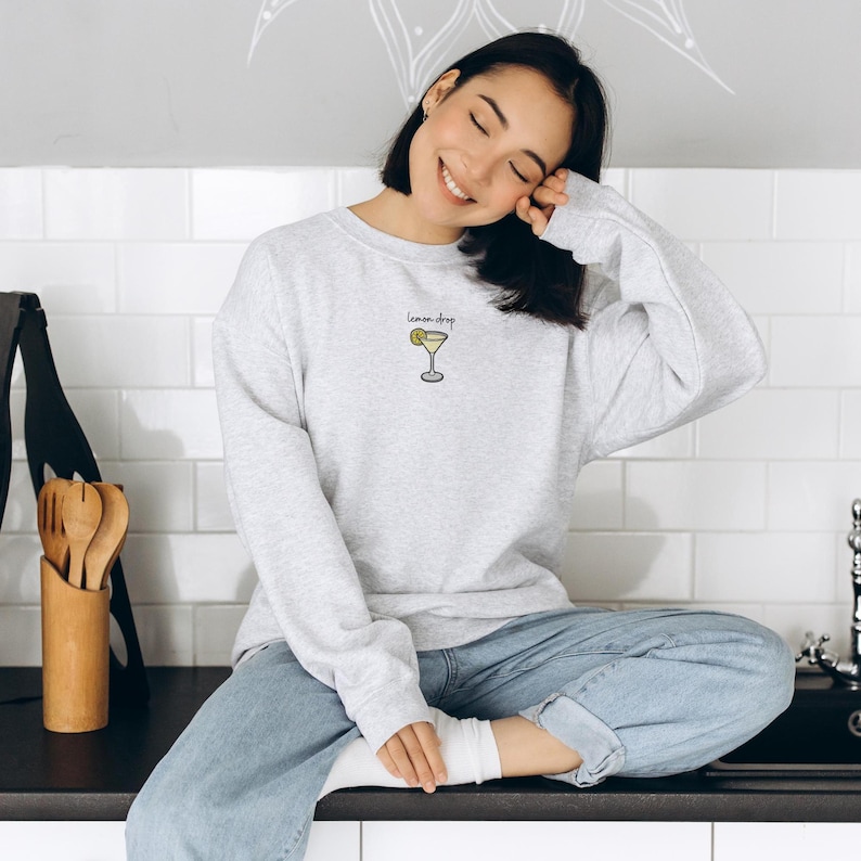Pu&ograve; includere: Una felpa grigia con la scritta "lemon drop" e un disegno di un bicchiere da cocktail con una fetta di limone. La modella indossa jeans blu e siede su un bancone.