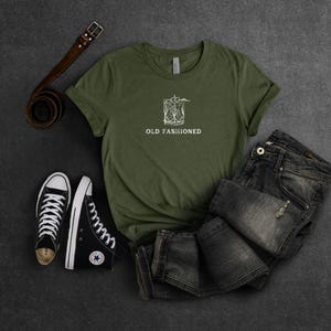 May include: A green t-shirt with a white graphic of a cocktail glass and the text "Old Fashioned". The t-shirt is laid out on a gray surface with a pair of black sneakers and a brown leather belt.